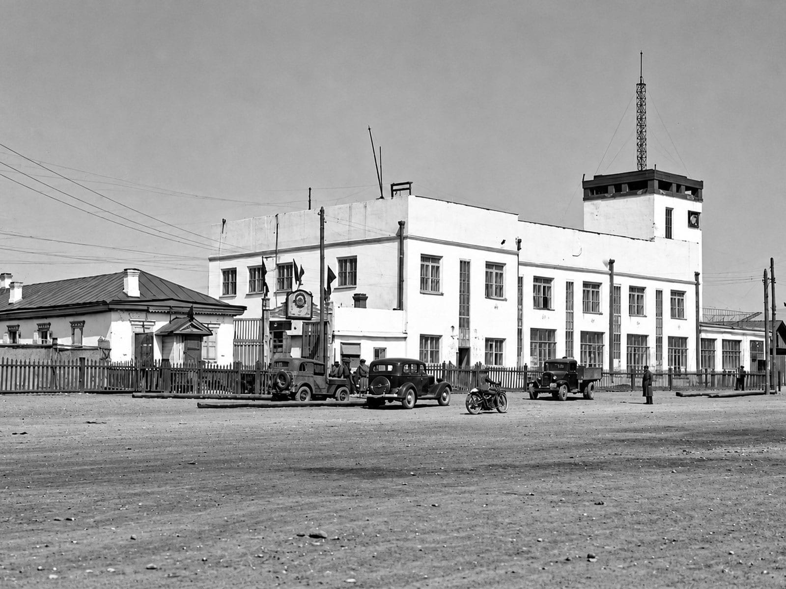 A white communications building with a tower and radio mast, and period vehicles in front.