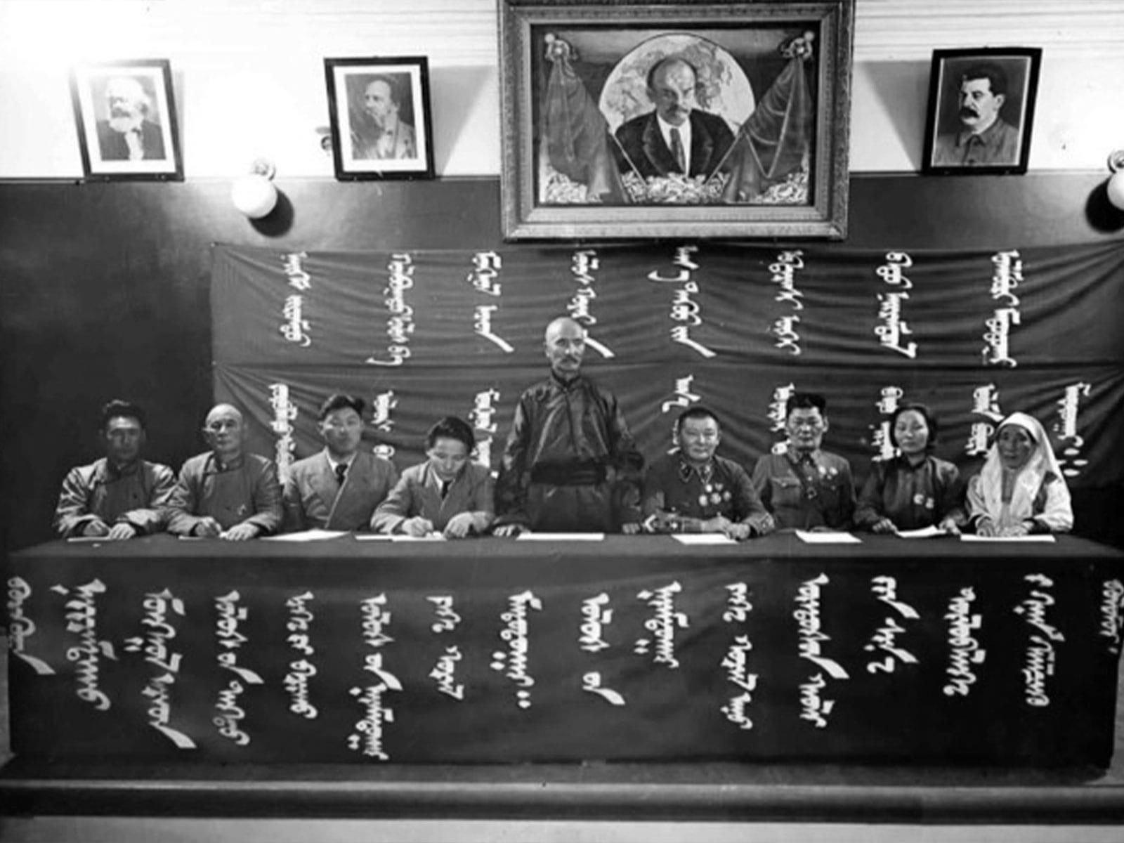 A formal meeting of communications officials beneath portraits.