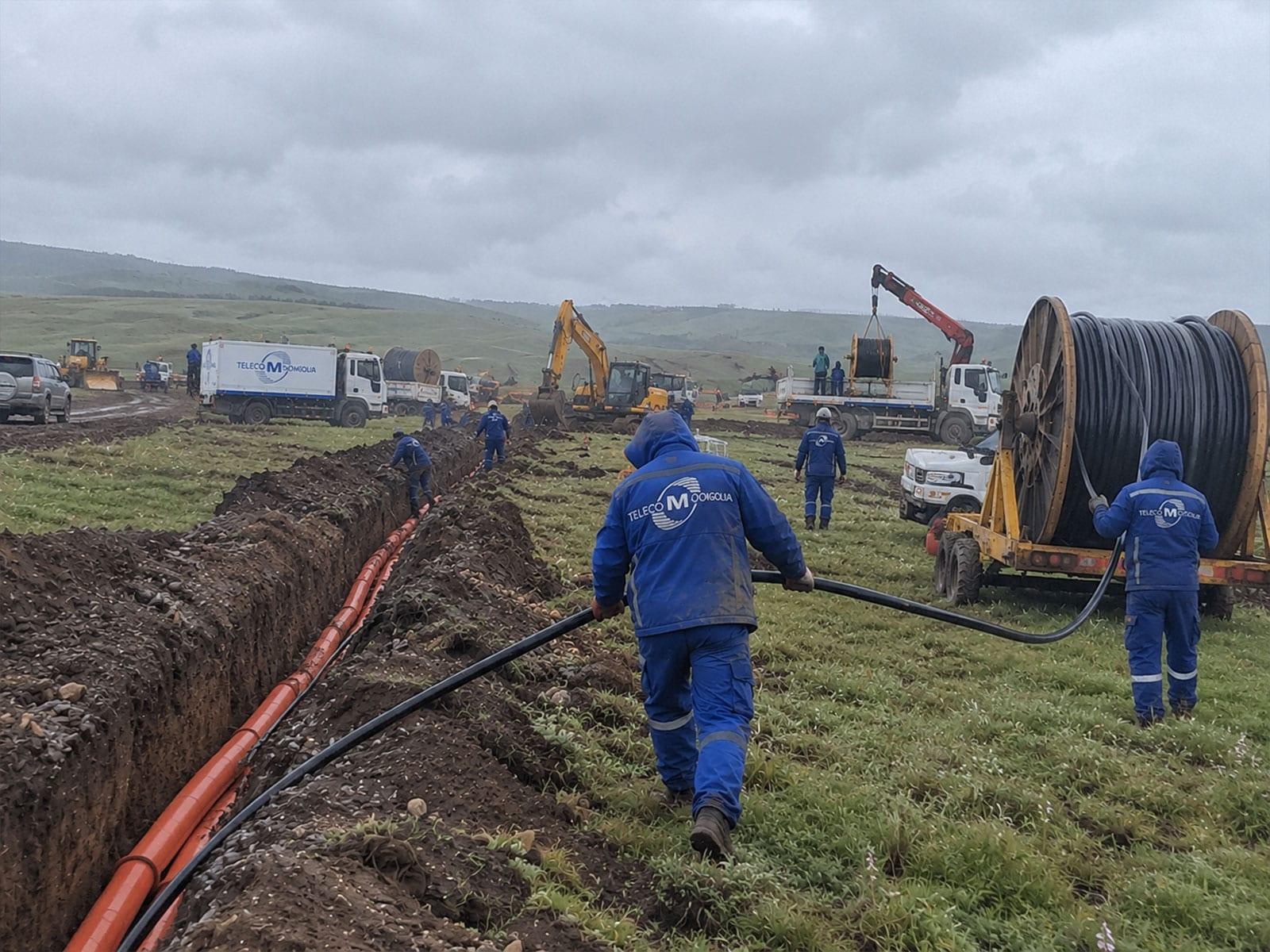 Crews in blue workwear laying cable across open country.